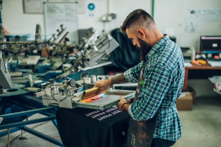 Male worker pressing ink on frame while using a printing machine in a workshop.